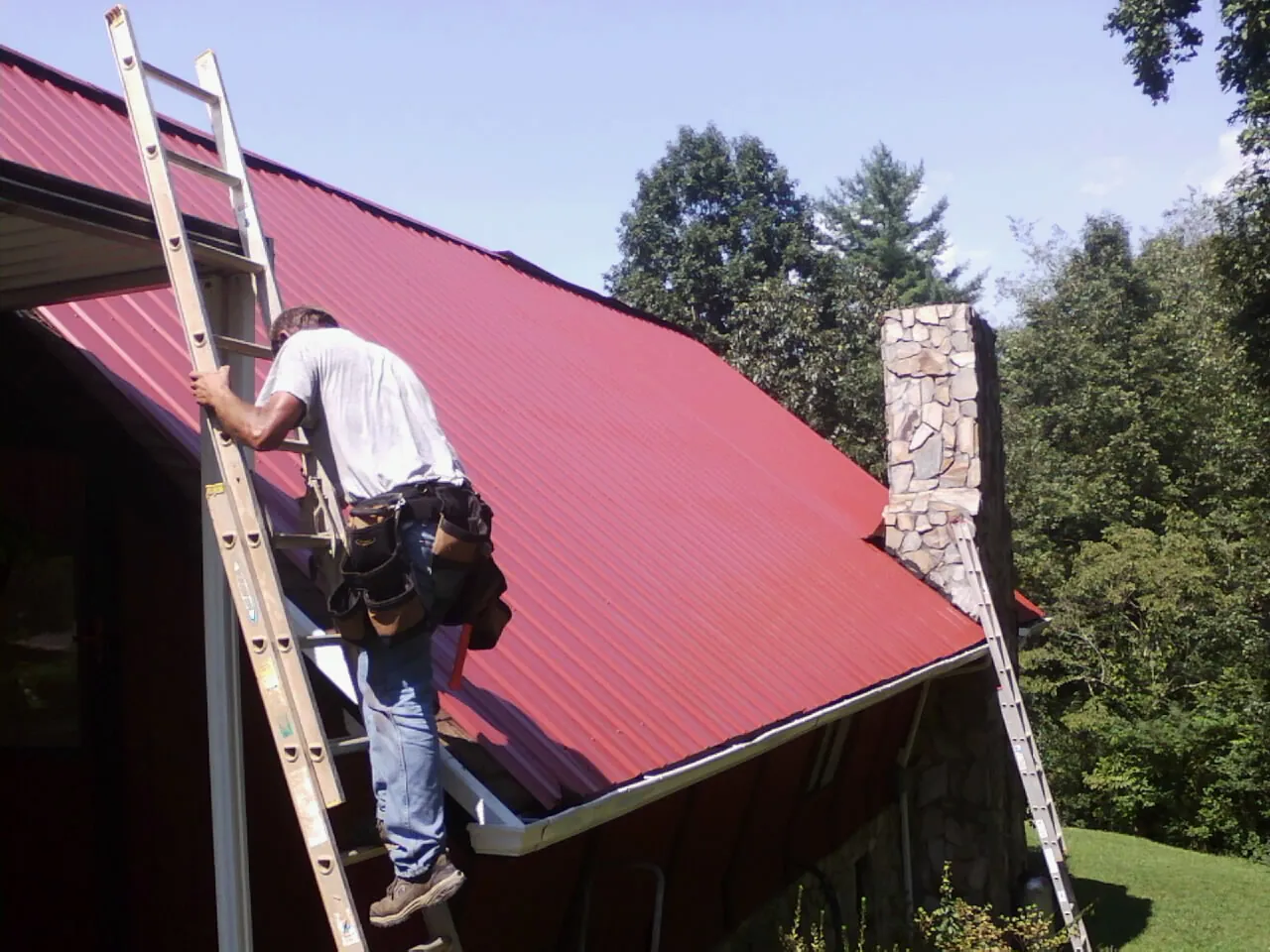 Roofer on a ladder beside a red metal roof and stone chimney — Soffit & Fascia Repair by Roofing Brothers in New Berlinville